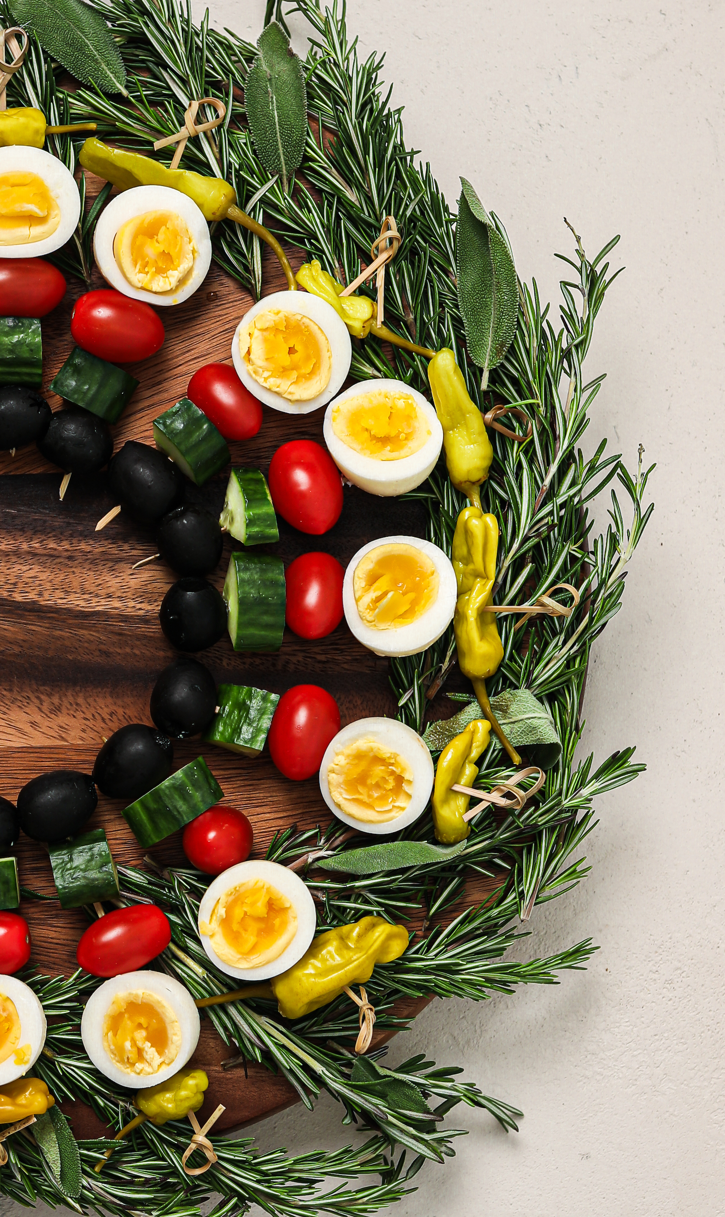 Overhead image of half a colourful and festive wreath of wooden picks threaded with boil egg halves, veggies and chilli peppers on a bed of rosemary and sage.