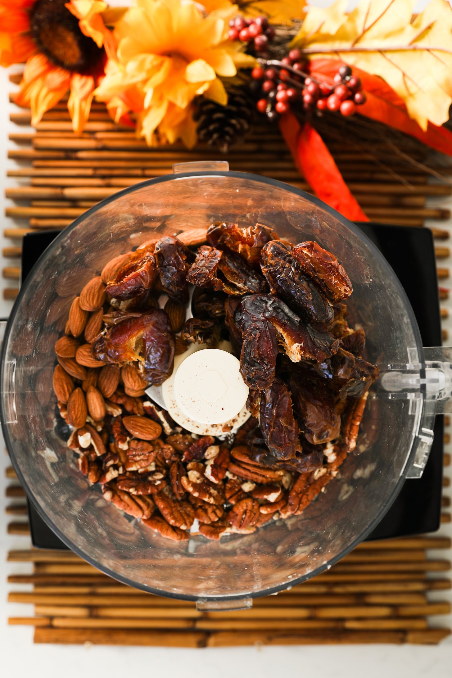 Top view of a food processor filled with dates, almonds and pecans, with seasonal flowers positioned nearby.