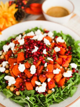 Perspective image showing a layered salad with a bed of arugula, roasted pumpkin pieces, pomegranate kernels, seeds, and chunks of goat cheese on top.