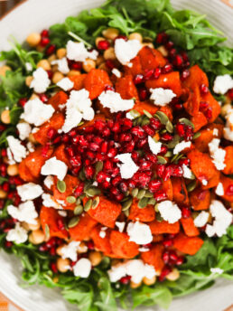 Overhead close up image featuring a layered salad with arugula roasted pumpkin pieces, pomegranate kernels, seeds, and chunks of goat cheese on top.