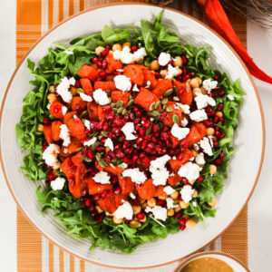 Overhead image featuring a layered salad with arugula roasted pumpkin pieces, pomegranate kernels, seeds, and chunks of goat cheese on top.