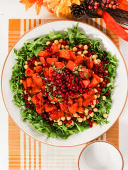 A large plate with a bed of arugula, chickpeas, roasted pumpkin pieces, pomegranate kernels and pumpkin seeds set on a placemat with orange and yellow flowers nearby.