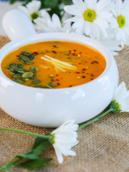 An angled view of an orange-coloured soup in a bowl, crowned with sunflower seeds, ginger strips, and chilli flakes, set against a backdrop of white flowers.