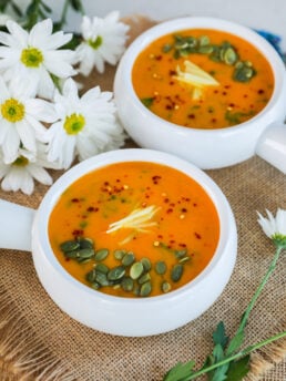 A perspective shot capturing two bowls of vibrant orange-hued soup adorned with seeds, ginger strips, and chilli flakes, complemented by white flowers in the background.