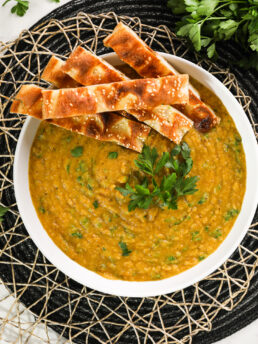 Close up image of a large bowl of creamy soup garnished with fresh parsley leaves and naan strips.