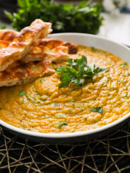 Perspective image of a large bowl of creamy soup topped with parsley leaves and naan strips.