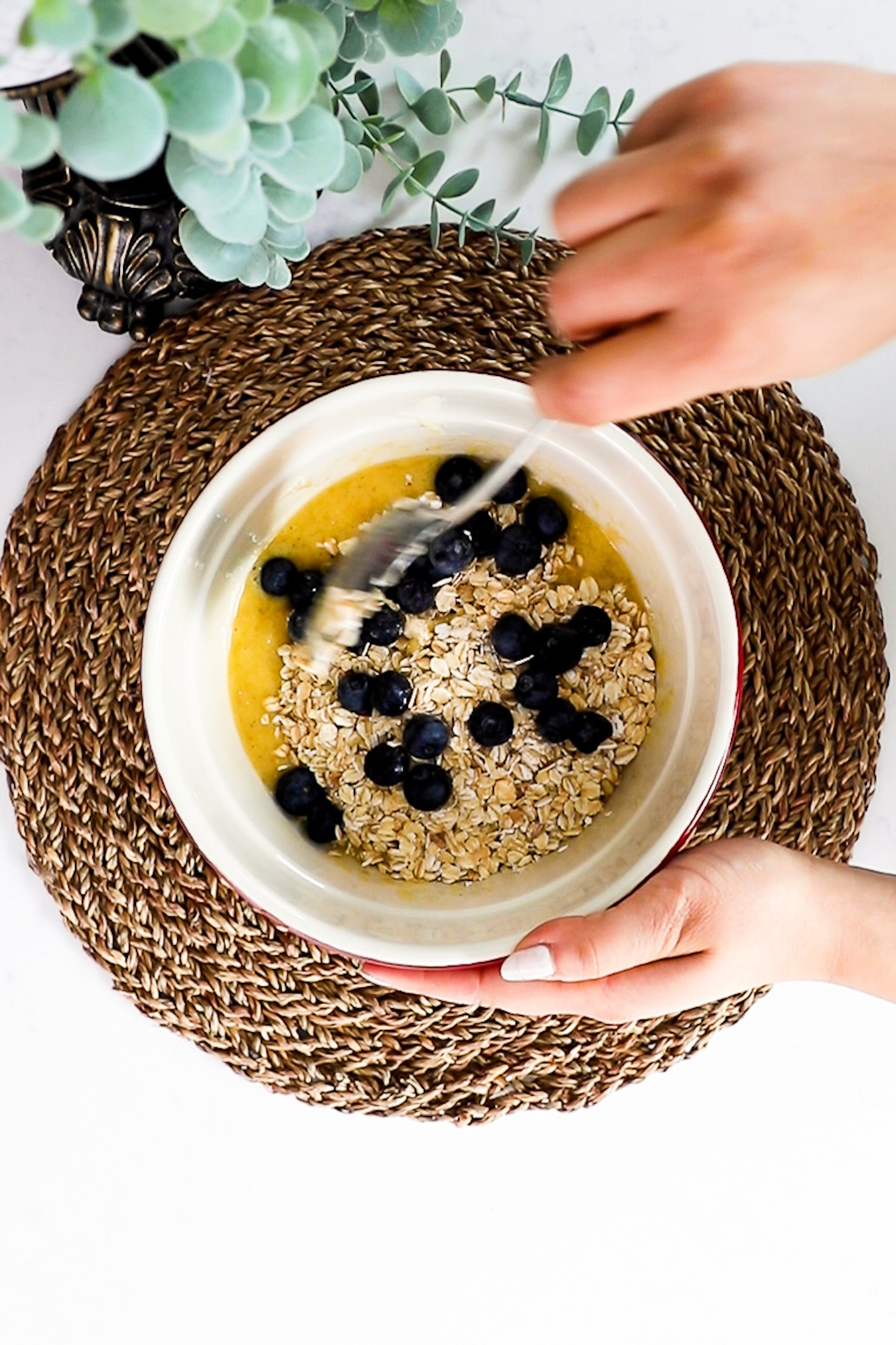 A hand holding a bowl and spoon mixing oats and blueberries into the wet ingredients.