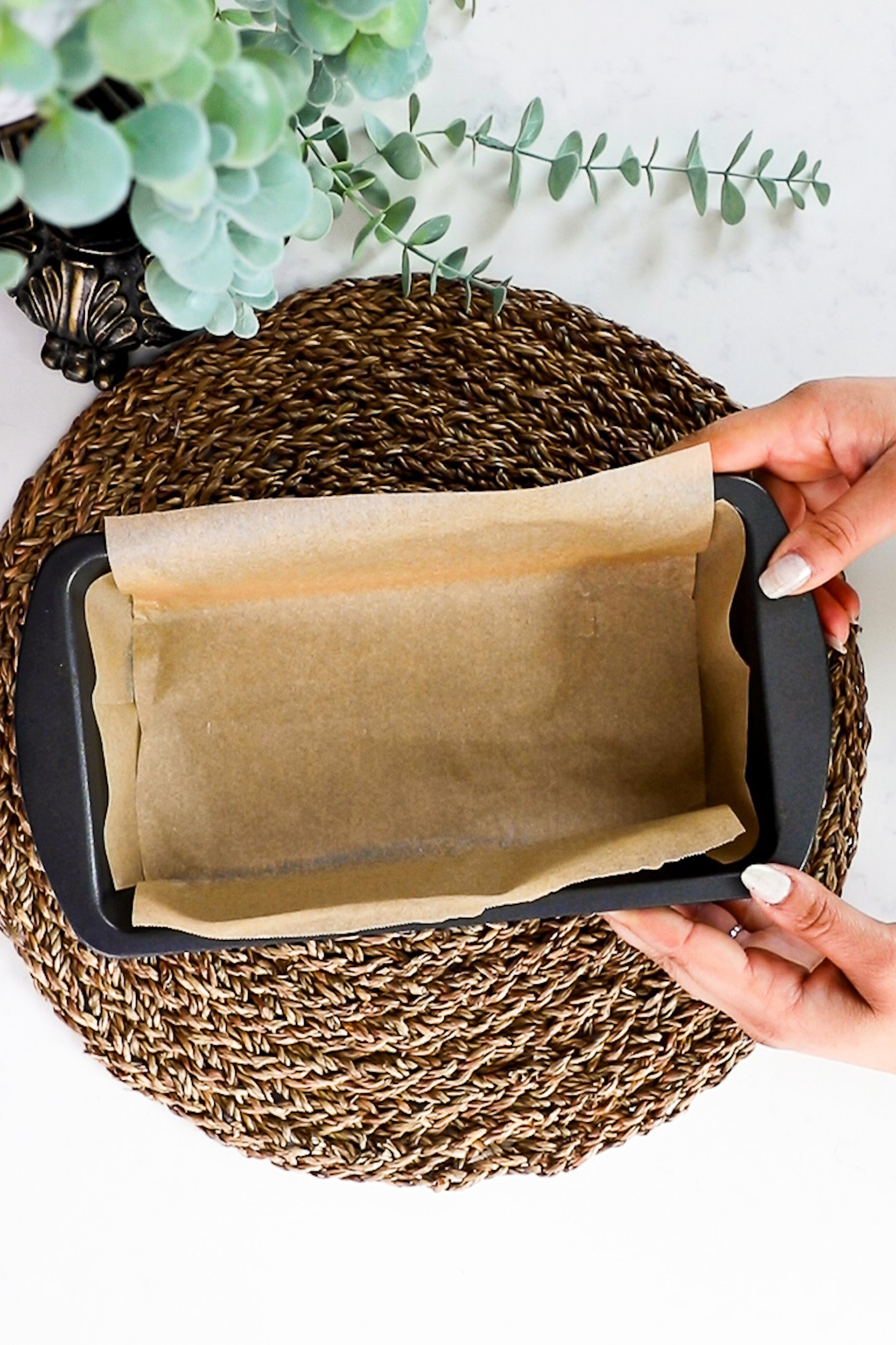 A hand holding a loaf pan lined with parchment paper on a round straw mat.