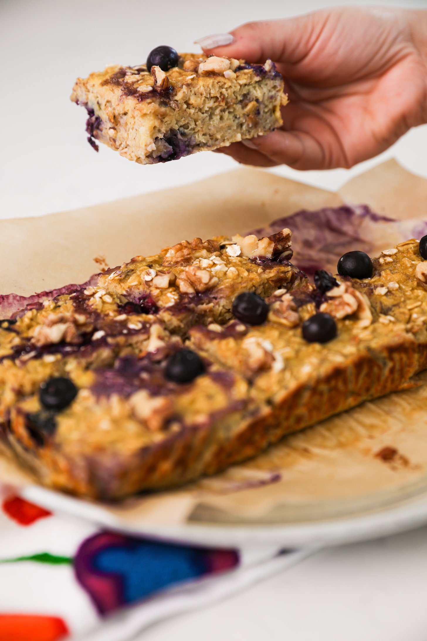 Hand holding a baked oatmeal bar over a plate with five more squares topped with blueberries.
