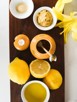 A display of food ingredients showing lemon, honey, mustard, oil and garlic on a dark wooden board.