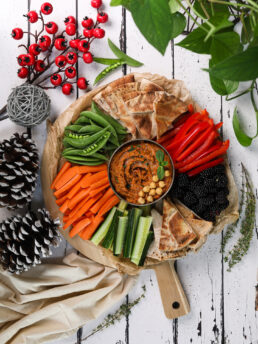 Overhead image of a vibrant platter of veggies, berries, pita and hummus styled with a holiday theme.
