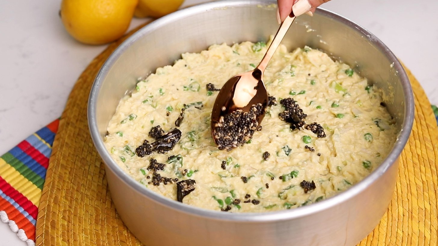 Hand uses a spoon to drizzle an oily seeded mixture onto the vegetable-semolina-yogurt mixture.
