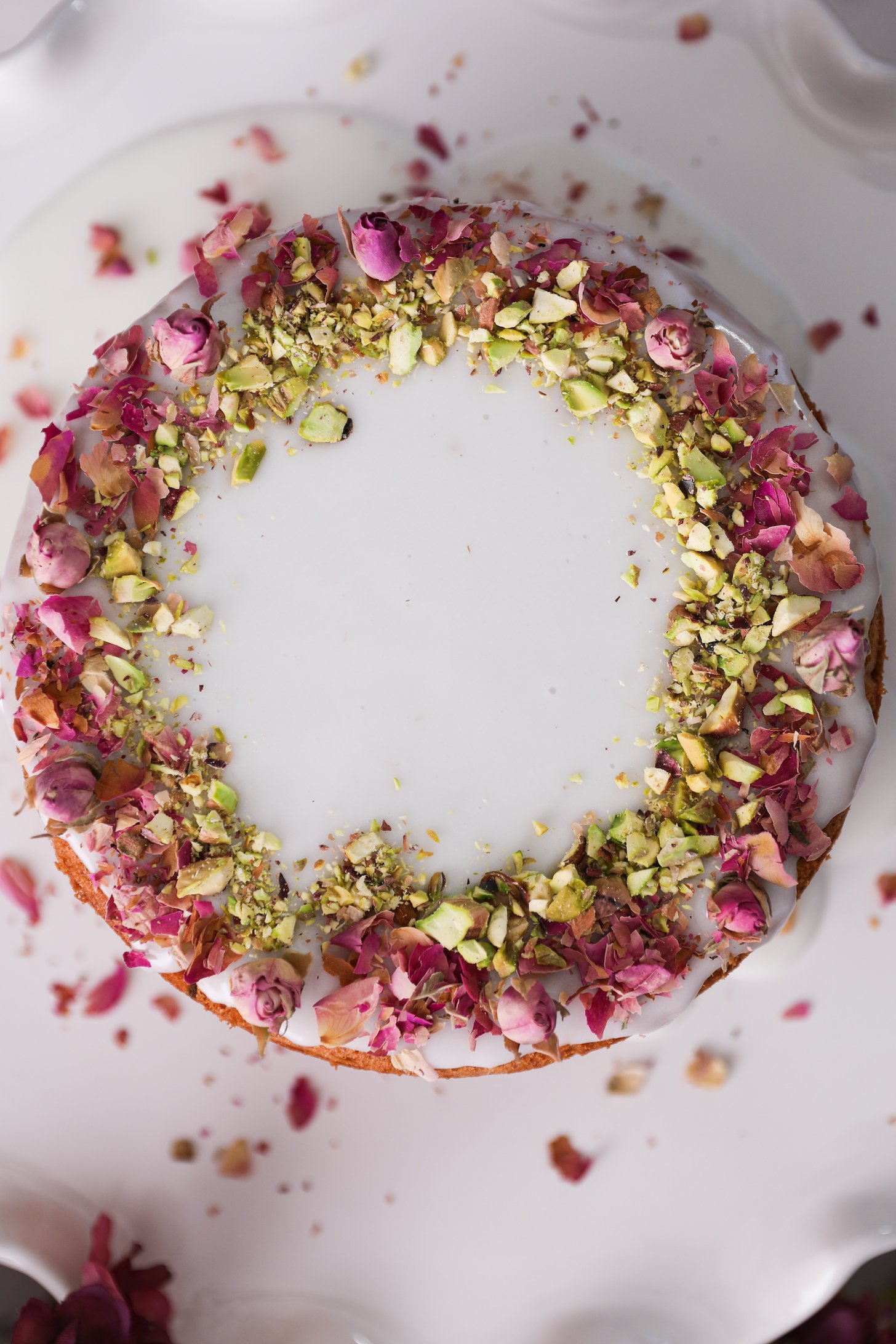 A white cake stand holds a round cake topped with smooth white icing with a boarder of red roses and crushed pistachios.