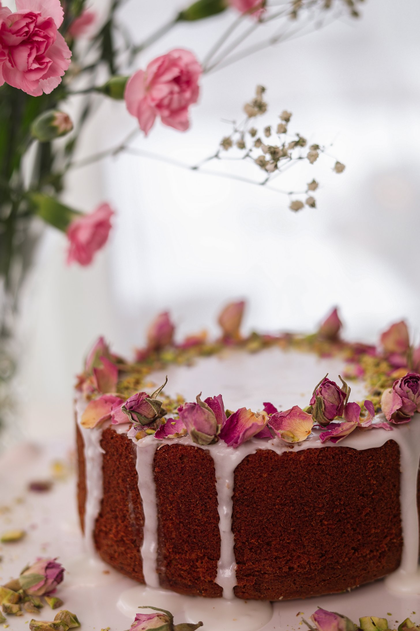 A perspective image of a cake with dripping white icing, dried roses, and crushed pistachios on top. Pink flowers lean over the cake.