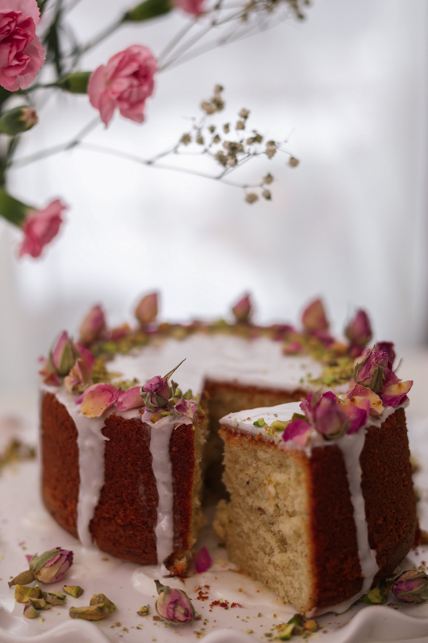 A perspective image of a cake with dripping white icing, dried roses, and crushed pistachios on top. A slice is pulled out and there are pink flowers lean over the cake.