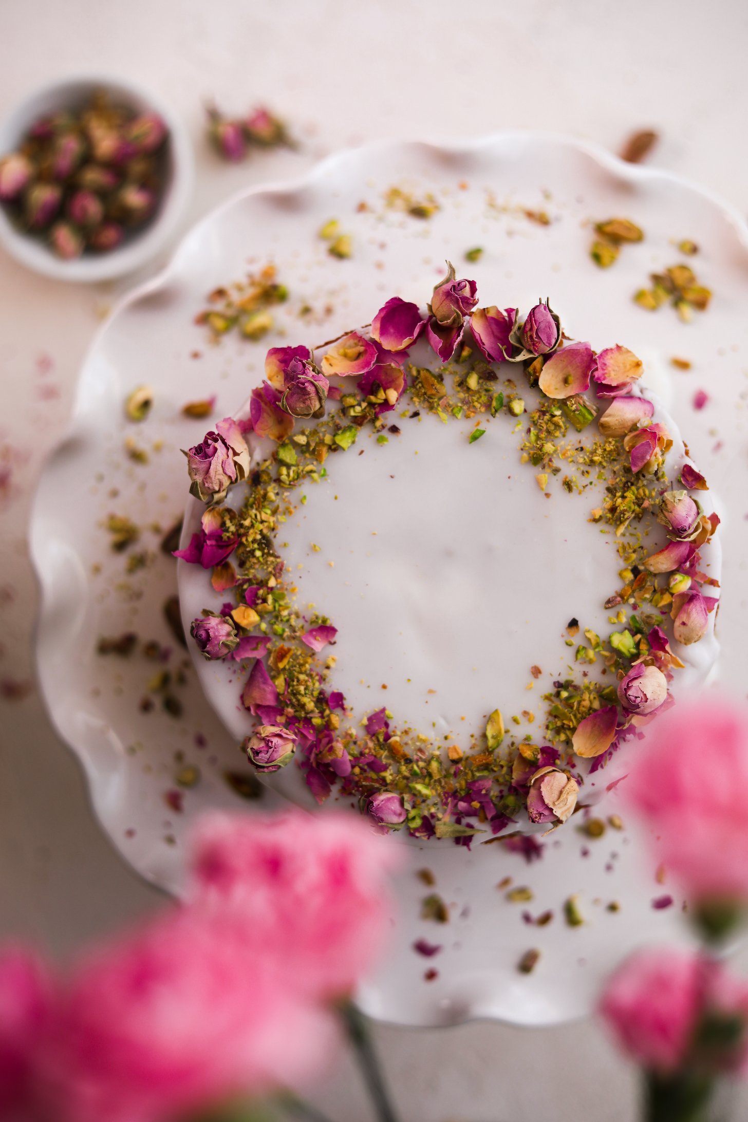From above, a cake stand holds a cake with white icing, dried roses, and crushed pistachios. Pink flowers lean over the cake.