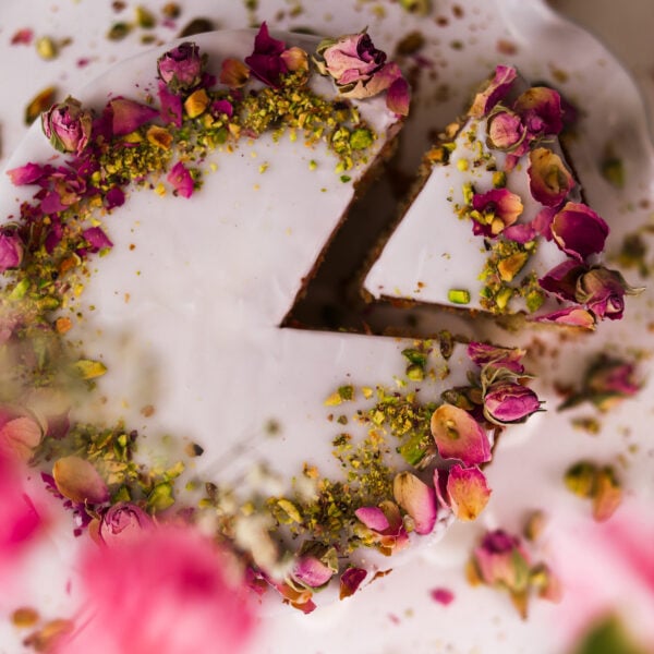 The top-down snapshot displays a cake on a cake stand. It's topped with white icing, dried roses, and crushed pistachios on top. A slice is pulled out.