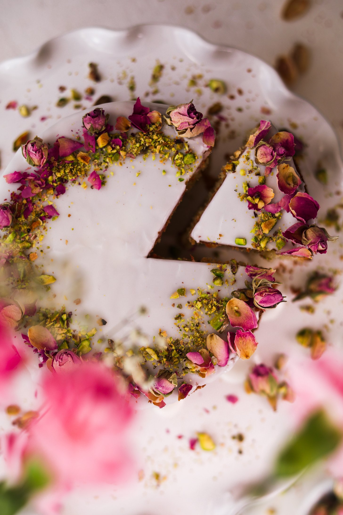 The top-down snapshot displays a cake on a cake stand. It's topped with white icing, dried roses, and crushed pistachios on top. A slice is pulled out.