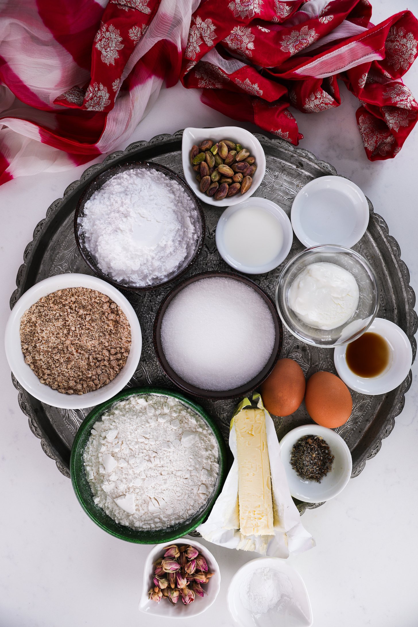 On a round tray, assorted food ingredients—flour, sugars, nuts, eggs, butter, dried roses, and spices—are neatly arranged. A traditional pink scarf rests nearby.