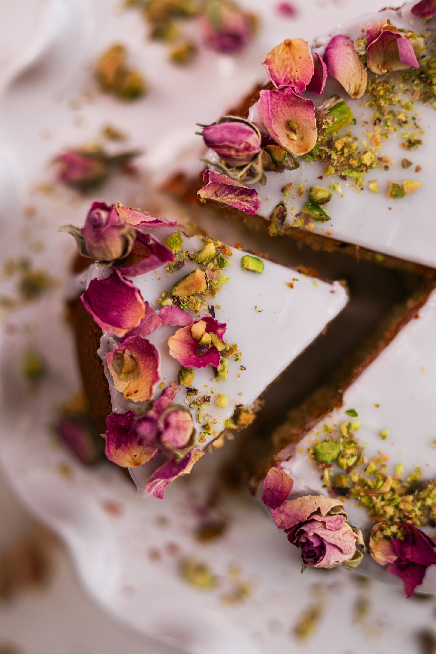 The close up top-down snapshot displays a cake featuring white icing, dried roses, and crushed pistachios on top. A slice is pulled out.