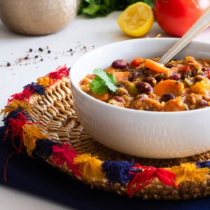 Perspective image of a bowl of vegetable chili on a colourful straw plate with vegetables and herbs in the background.