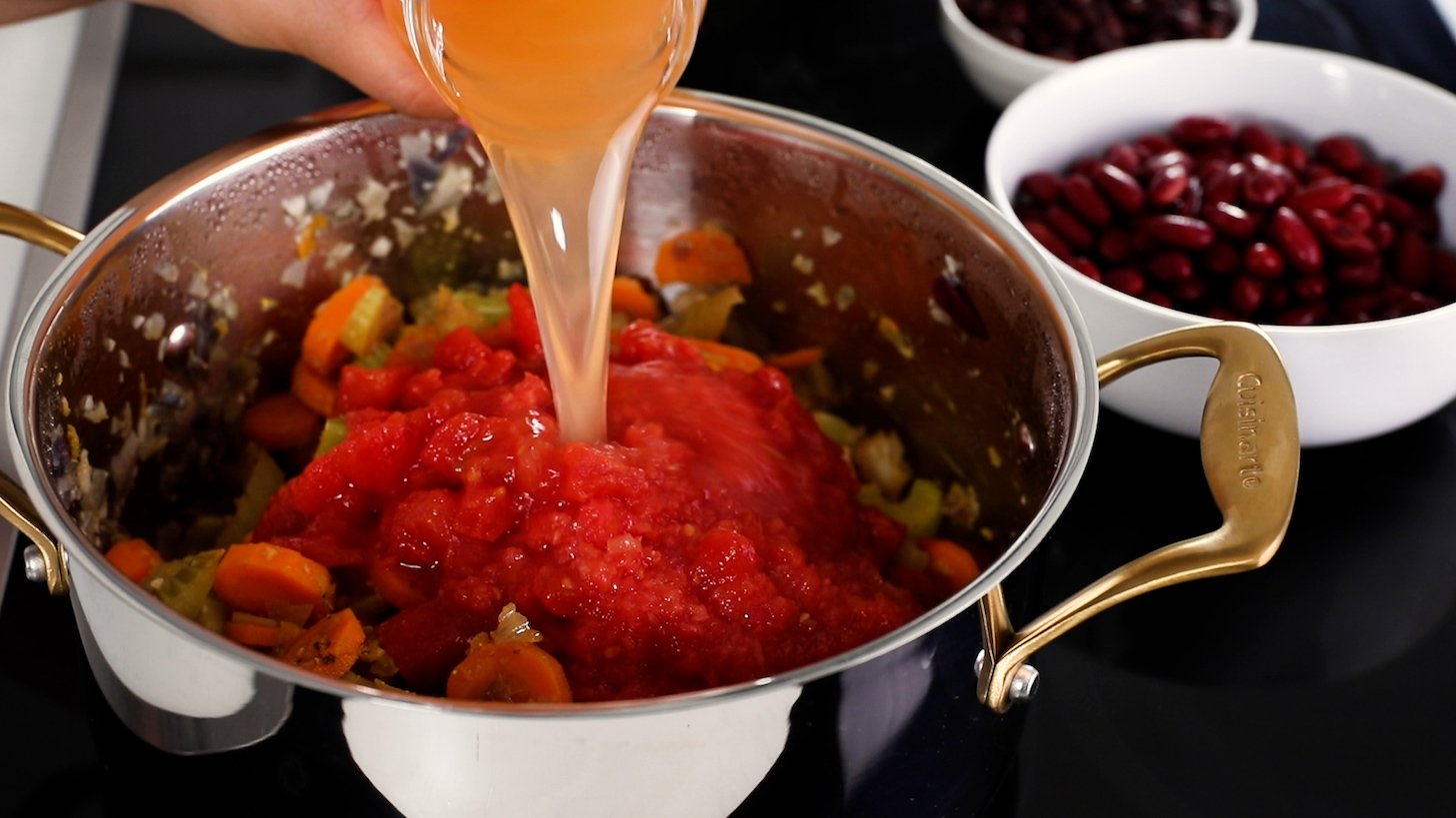A hand holding a jar of stock pouring it into a cooking pot filled with tomato puree and assorted vegetables.