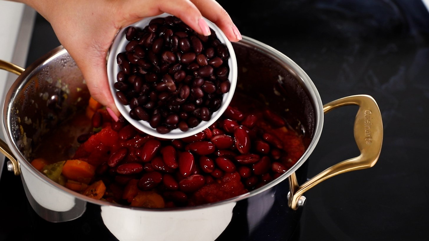 A hand holding a bowl of cooked black beans tipping it into a cooking pot filled with kidney beans and assorted vegetables.