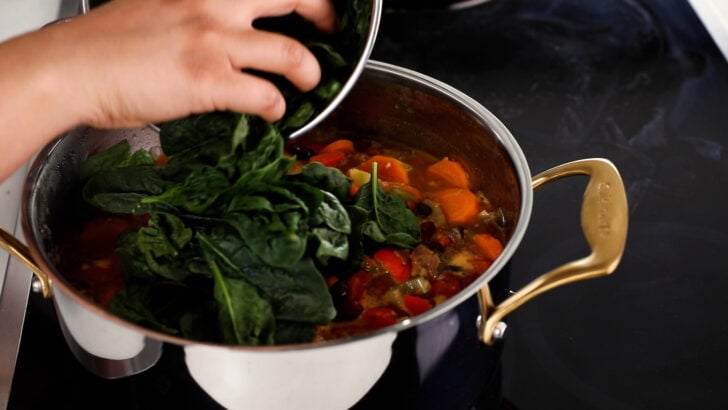A hand holding a bowl of fresh spinach, adding it into a cooking pot filled with assorted vegetables and stock.