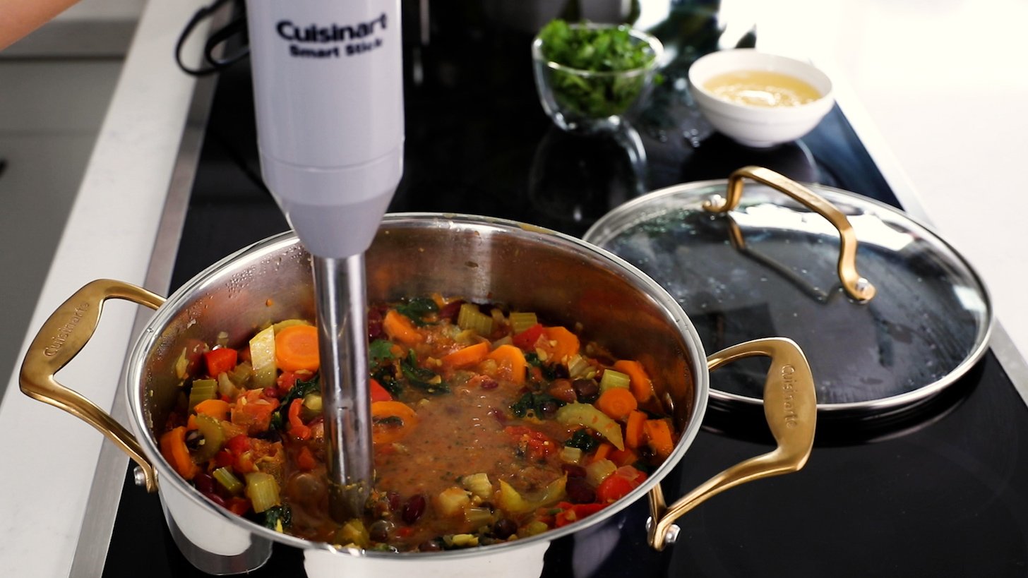 A hand-held immersion blender inside a cooking pot filled with an assortment of veggies and stock.