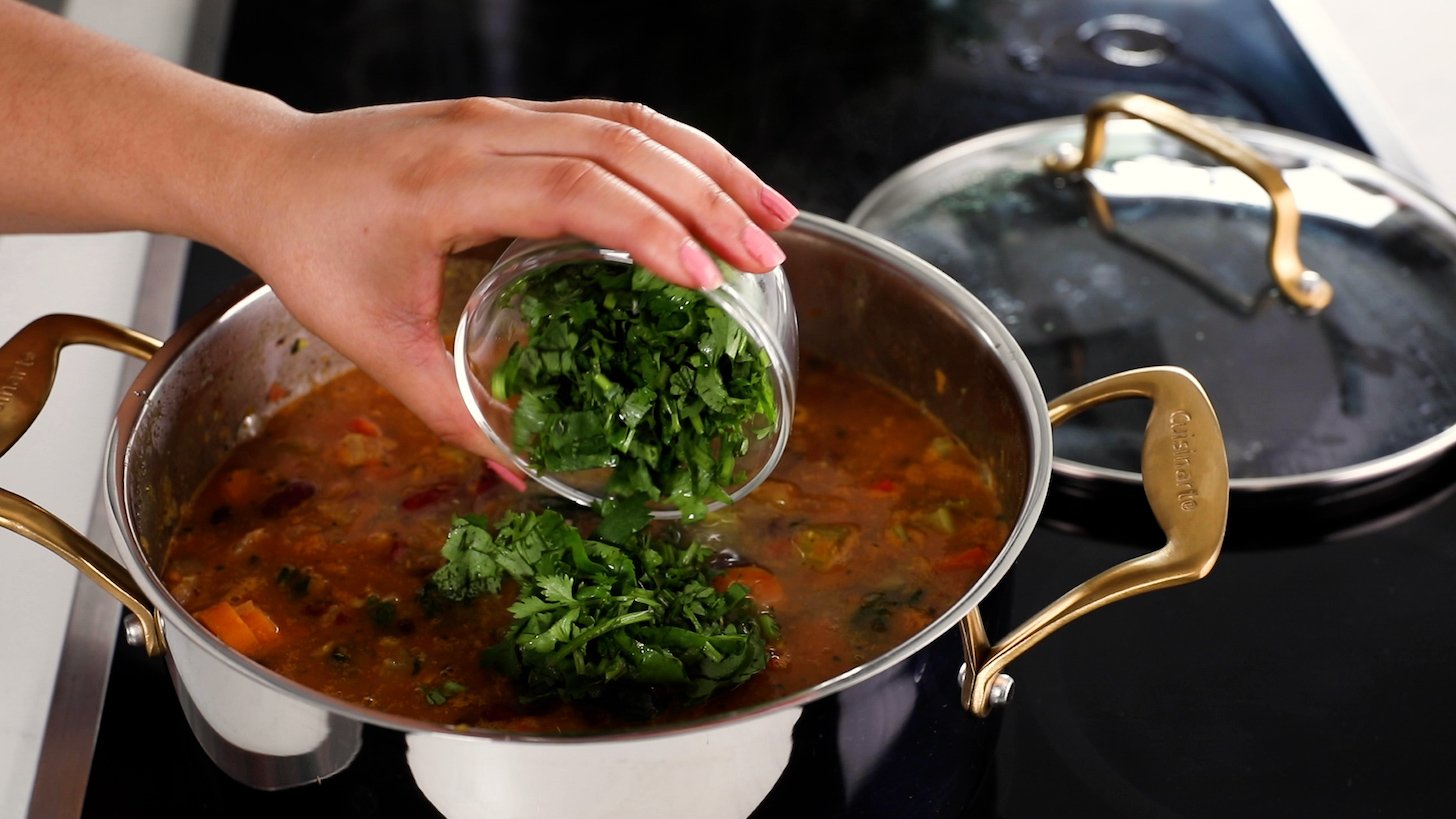 A hand holding a ramekin of chopped cilantro, adding it into a cooking pot full of chili.