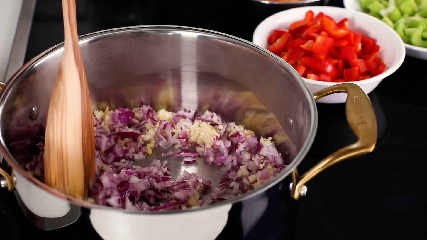 Chopped red onion and garlic placed in a cooking pot, a wooden spoon resting inside.