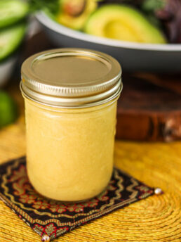 Perspective image of a mason jar of honey mustard dressing on a decorative mat with avocados in the bacground.