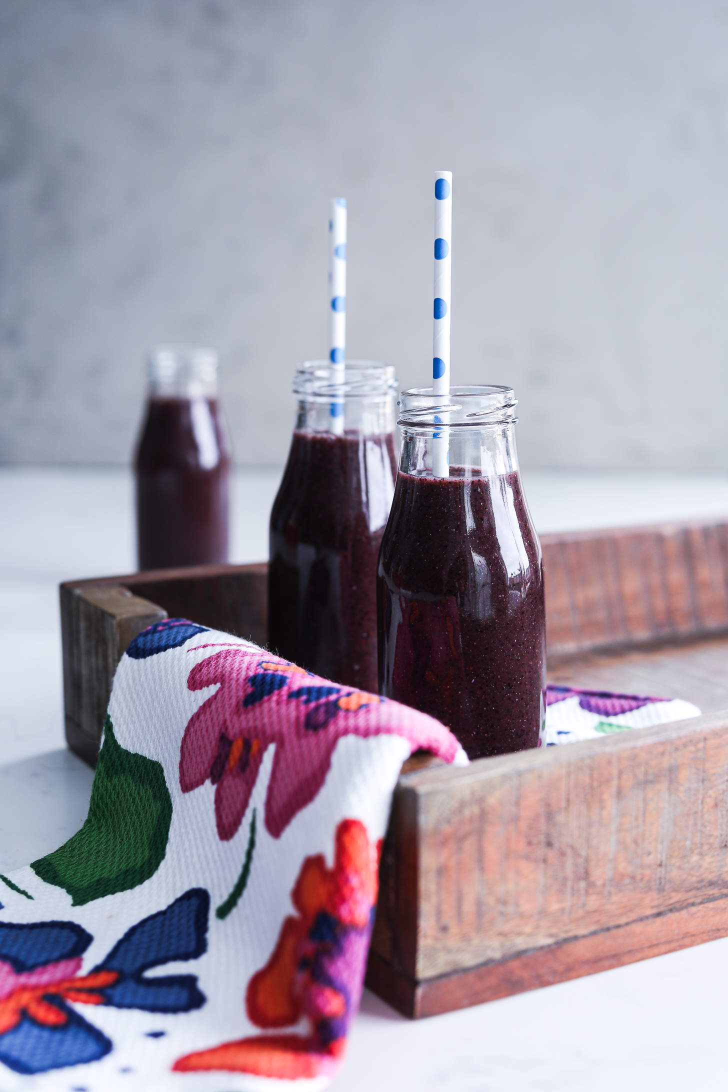 Blueberry kale smoothie in small bottles with straws on a tray, covered by a cloth extending out.