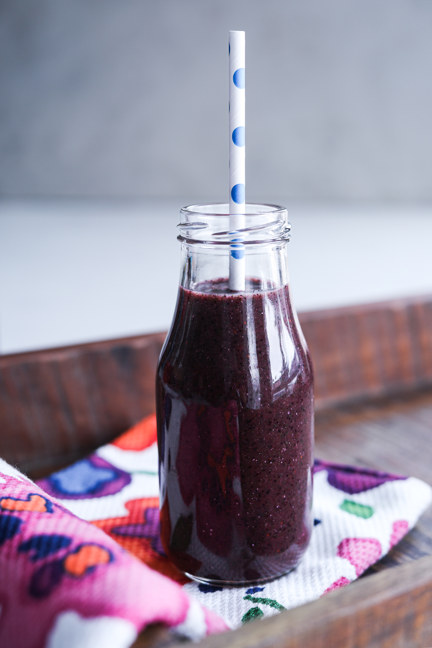 Close-up photo of a blueberry kale smoothie in a bottle with a straw, situated on a tray atop a cloth.