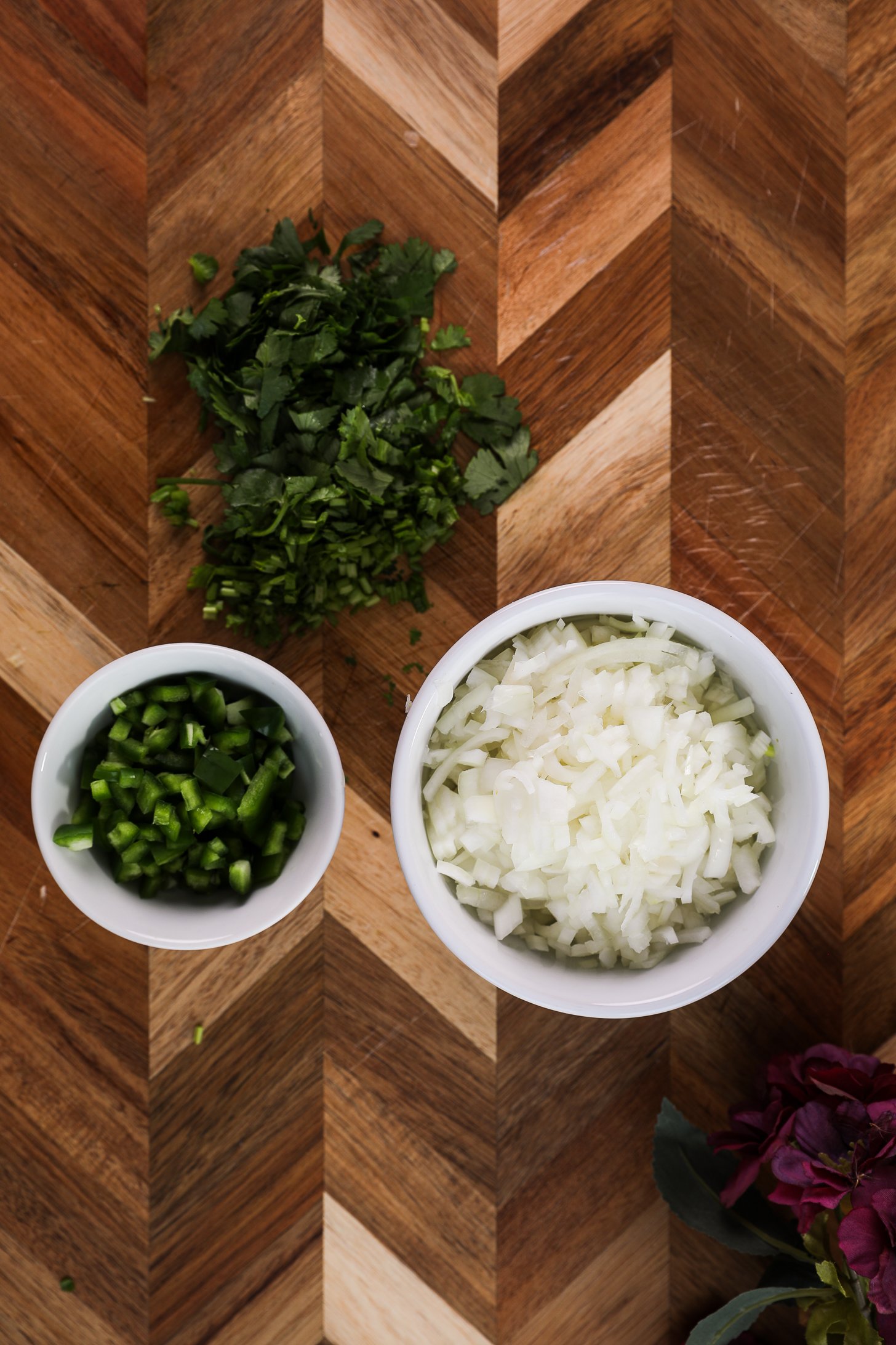 Wooden board featuring two ramekins of chopped onion and jalapeno, accompanied by chopped cilantro nearby.
