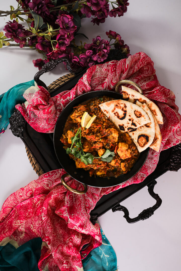 Bird's-eye view of an Indian wok with chicken, lentils, cilantro, lemon, naan, styled on a tray with dupatta scarf and nearby flowers.