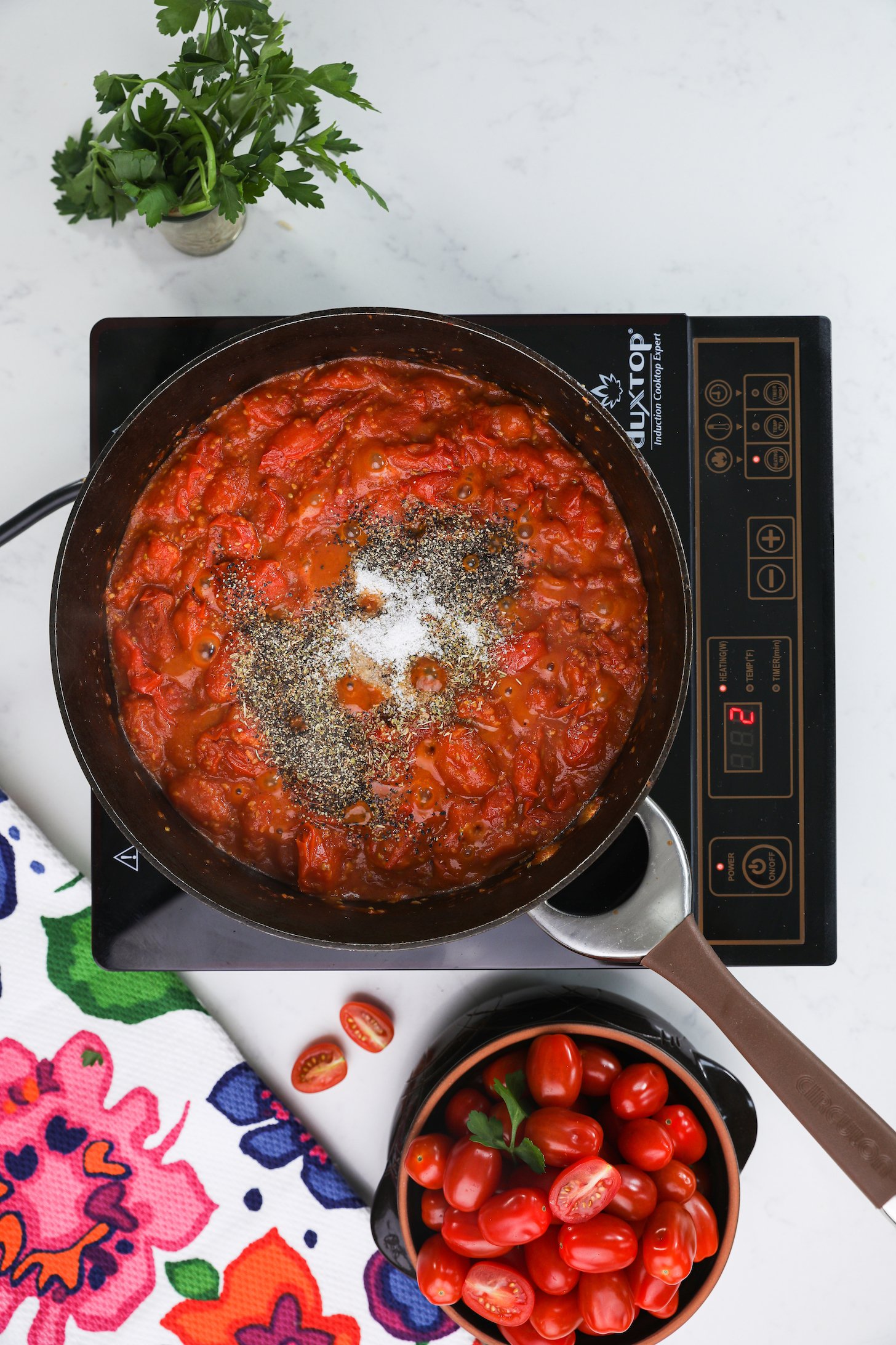 Tomato sauce topped with salt and pepper in a pan on a mobile cooktop, surrounded by herbs and fresh tomatoes.
