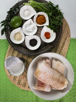 A selection of food ingredients includes, fish fillets, ramekins of spices and herbs as well as an open can of coconut milk.
