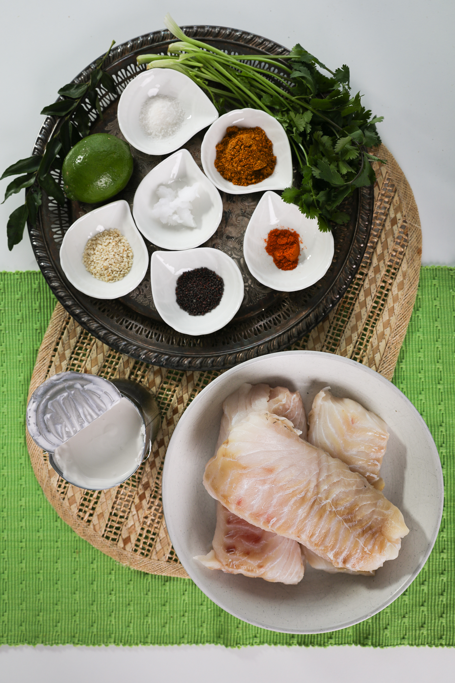 مجموعه ای از مواد غذایی شامل فیله ماهی، رامکین ادویه جات و گیاهان و همچنین یک قوطی باز شیر نارگیل است.