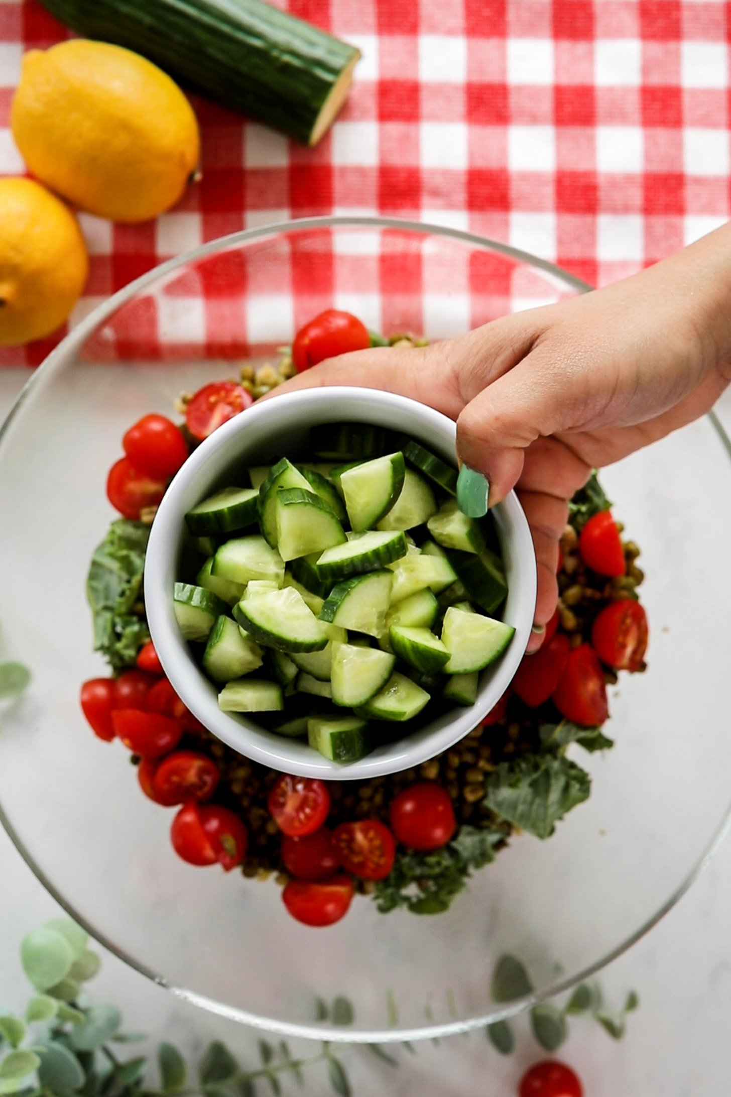 A hand holding a bowl of cucumber cubes over a salad bowl filled with cherry tomato halves and kale leaves.