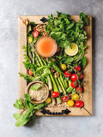 Variety of vegetarian healthy eating food ingredients. Green asparagus, herbs, tomatoes, nuts, wheat corns, dandelion leaves, glass of juice on wood tray over grey texture background. Top view, space.