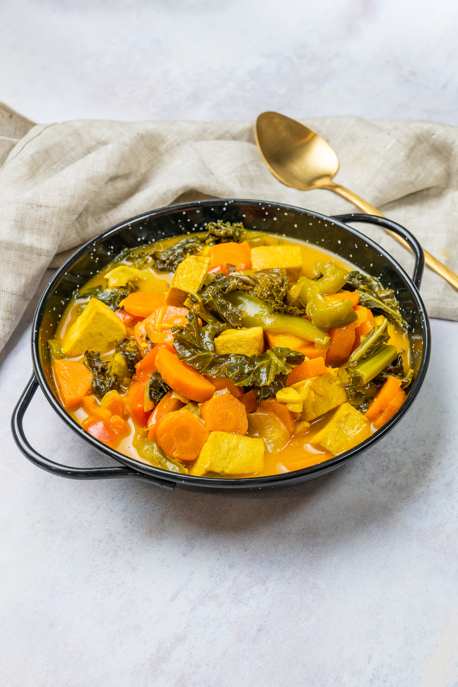Tofu, kale, and carrot soup in a shallow pan. It is located on a light gray background next to a golden spoon and a cloth napkin.