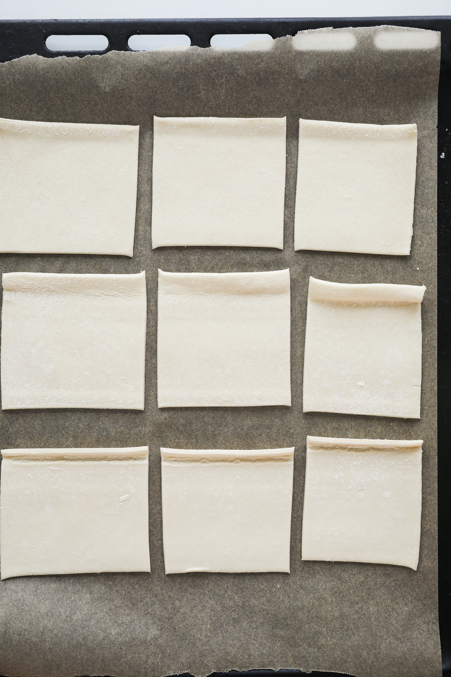 Top-down view of nine unbaked puff pastry squares arranged on a lined baking tray.