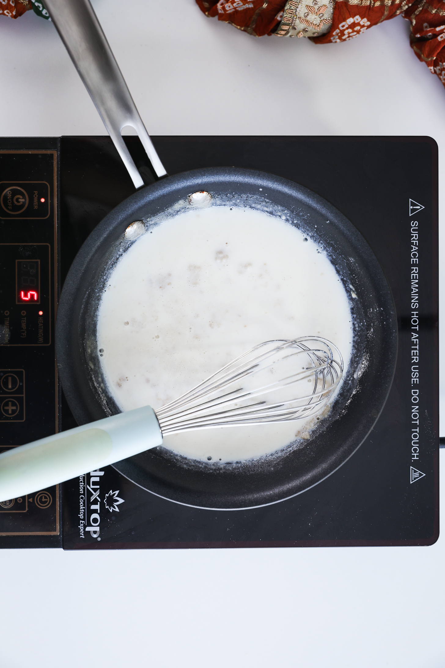 A pan filled flour and milk (making roux), featuring a whisk resting inside it, placed on a mobile cooktop.