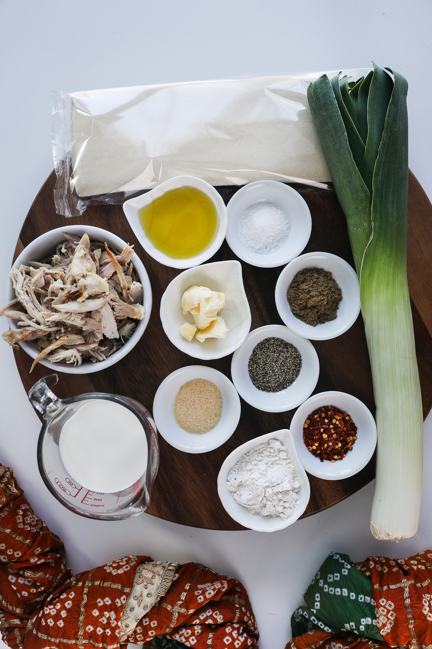 A collection of food ingredients including shredded chicken, leek, folded puff pastry, milk, flour and spices on a round board.
