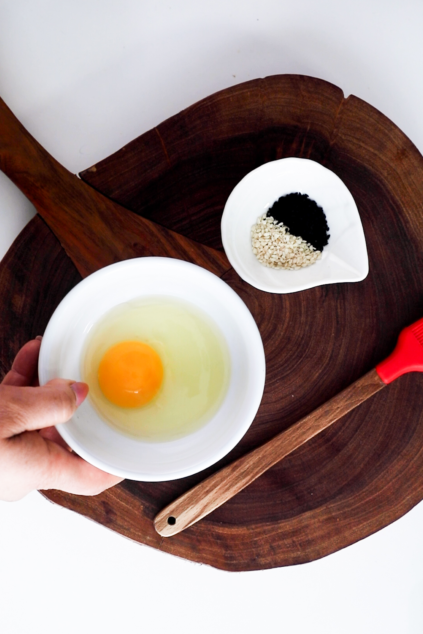 A hand holding a bowl with a cracked egg, accompanied by a ramekin of seeds, all arranged on a wooden board.