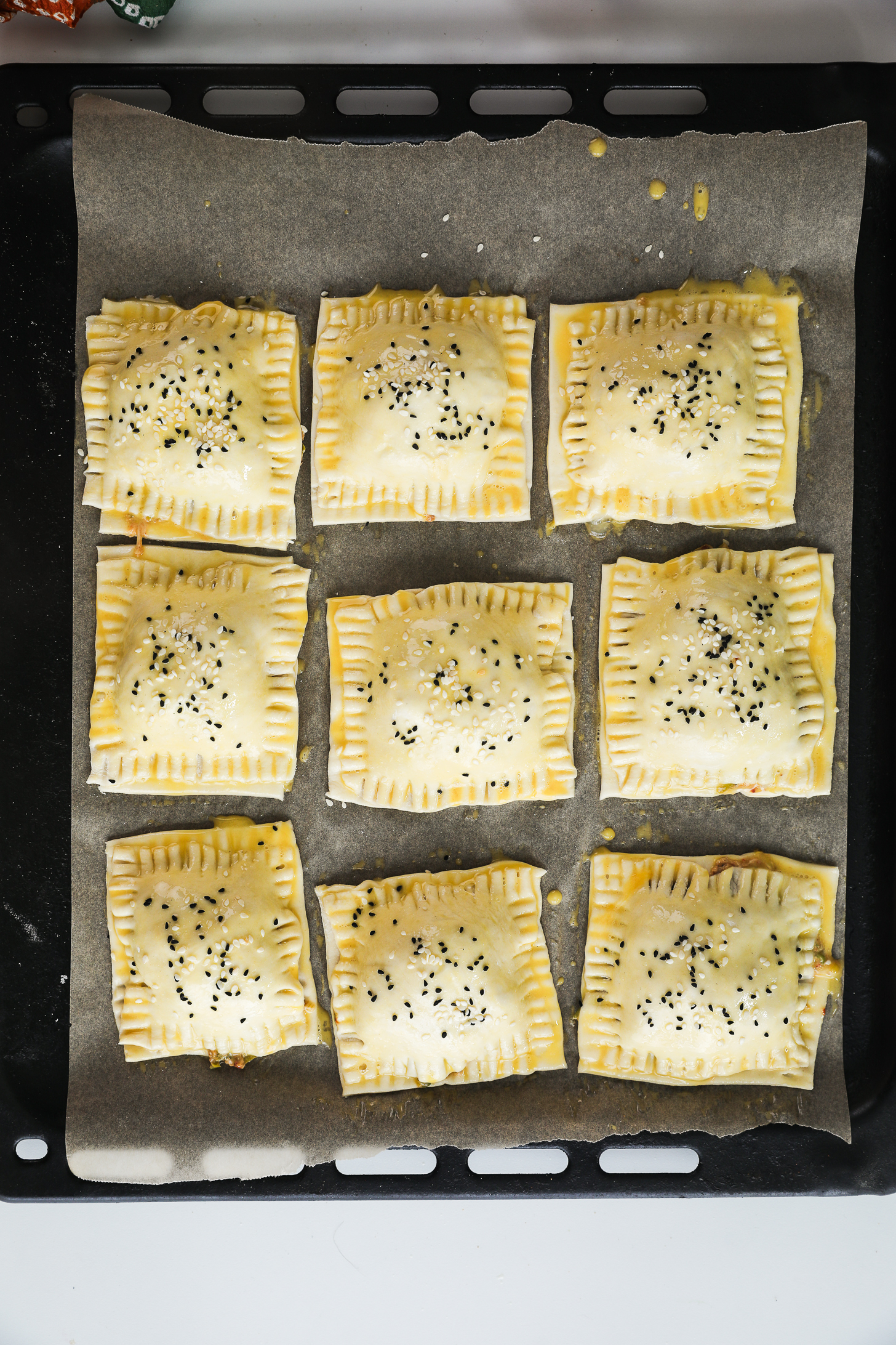 Top-down image of an oven tray lined with unbaked puff pastry squares, each filled with a filling, brushed with egg wash, and sprinkled with seeds.