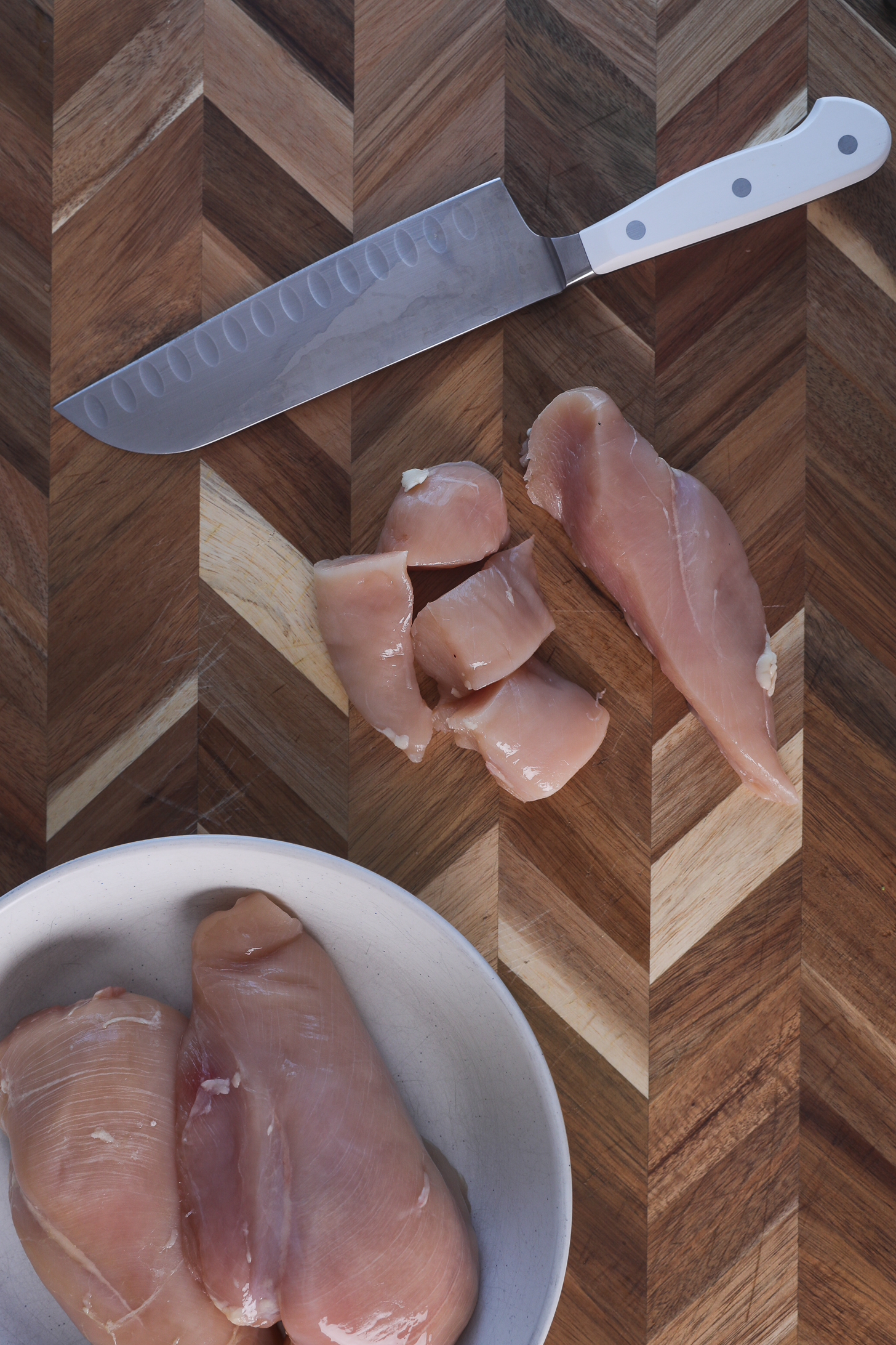 A chicken breast fillet half sliced into chunks on a wooden board with a nearby knife and a bowl of additional chicken breast fillets.