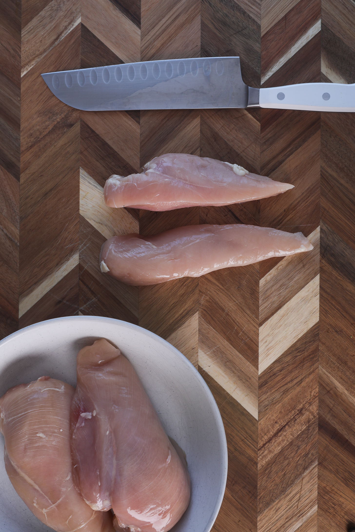 A chicken breast fillet sliced in half (lengthwise) on a wooden board with a nearby knife and a bowl of additional chicken breast fillets.