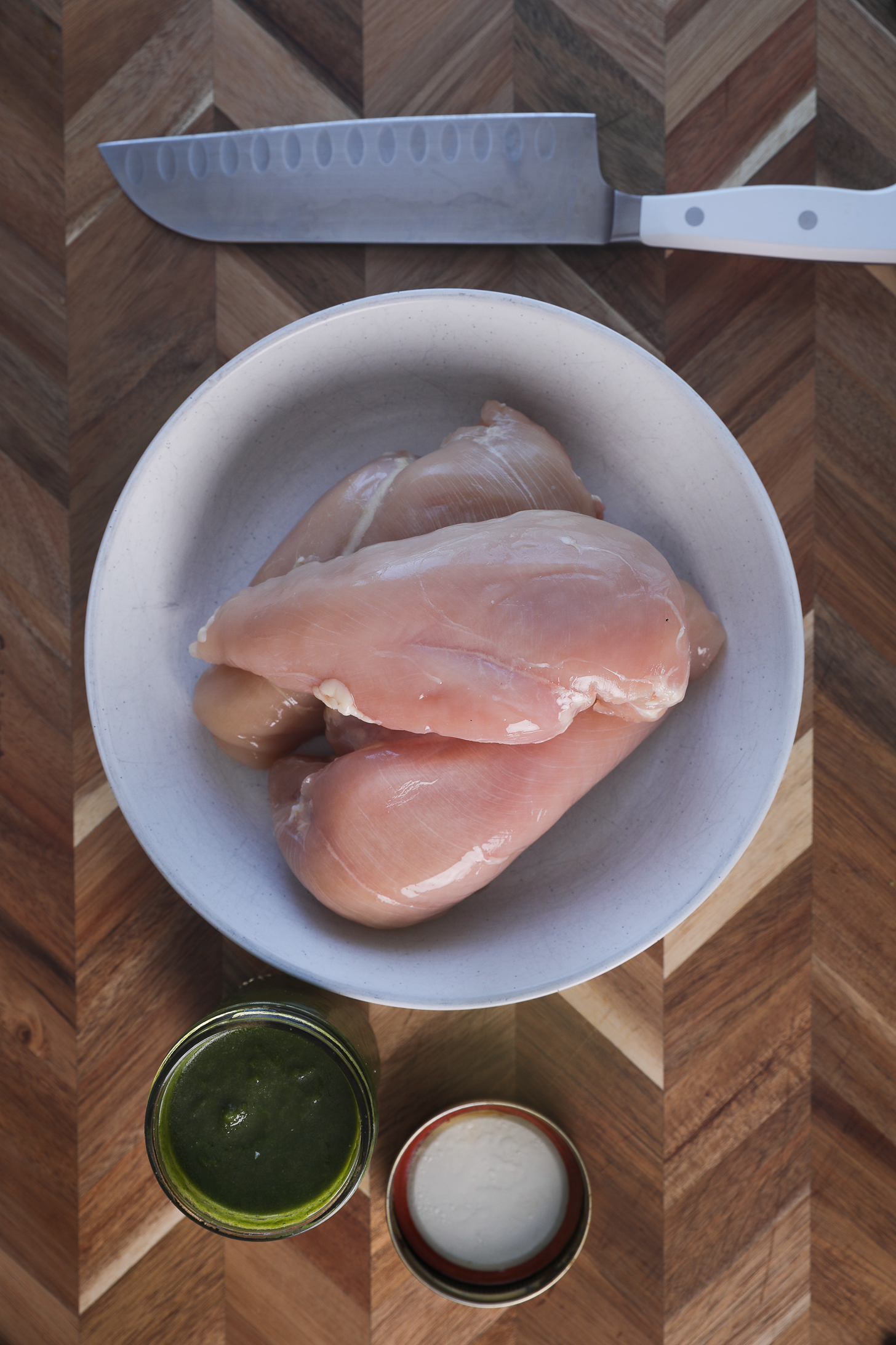 A bowl of chicken breast fillets placed on a wooden board next to a mason jar of green sauce.